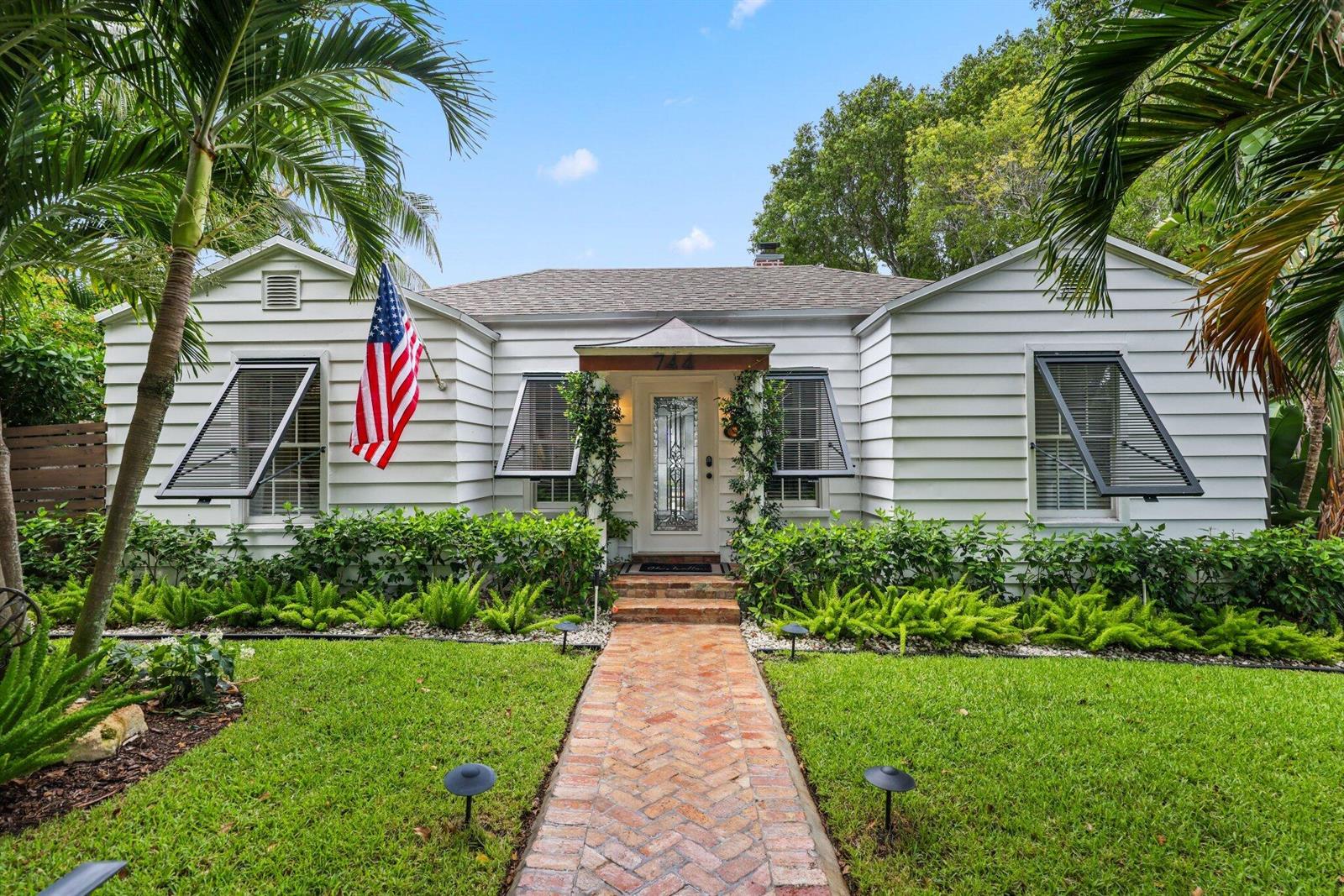 A white house with a brick walkway and an American flag stands behind a lawn — 744 Sunset Road, West Palm Beach FL 33401, WEST PALM BEACH, FL