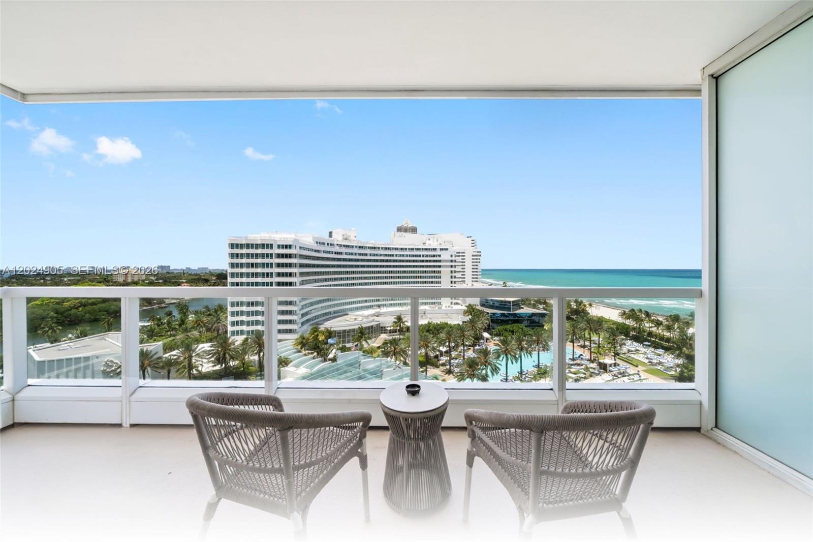 A balcony features two woven chairs and a small table overlooking a resort and ocean — 4391 Collins Ave # 1001, Miami Beach FL 33140, MIAMI BEACH, FL