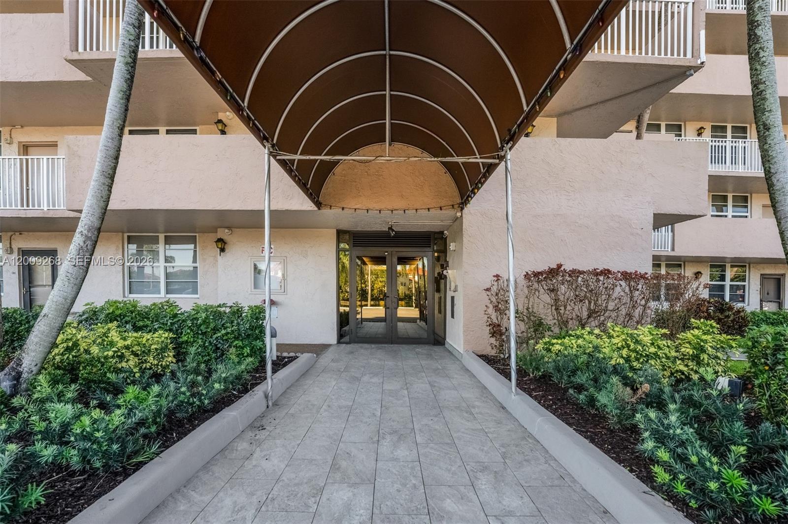 A paved walkway leads to the glass-doored entrance of a multi-story building under a canopy — 250 Jacaranda Dr # 508, Plantation FL 33324, PLANTATION, FL