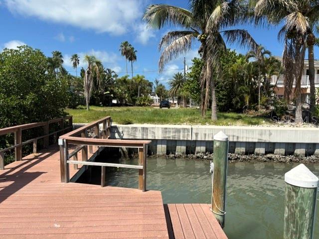 A wooden dock extends over a canal toward a grassy lot with palm trees — 14065 N BAYSHORE DRIVE, MADEIRA BEACH, FL