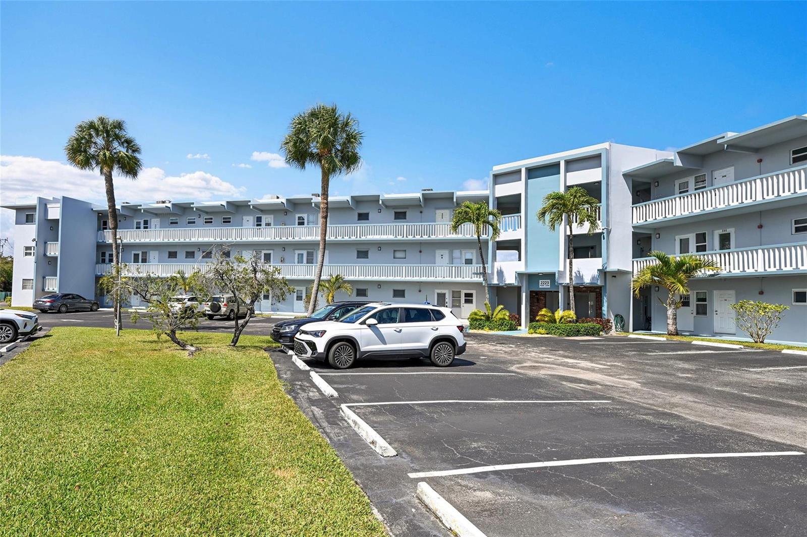 A multi-story apartment building with a parking lot and palm trees under a blue sky — 2223 Park Lane # 302, Hollywood FL 33021, HOLLYWOOD, FL