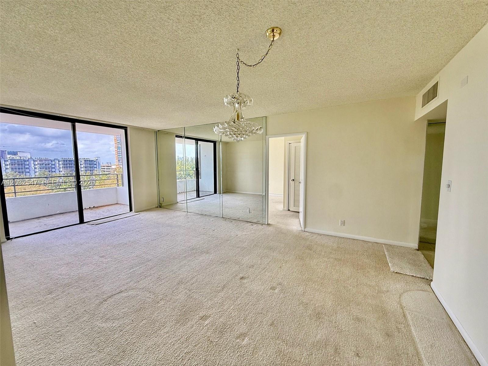 An empty living room features beige carpeting, a crystal chandelier, mirrored walls, and a balcony — 500 Three Islands Blvd # 314, Hallandale Beach FL 33009, HALLANDALE BEACH, FL