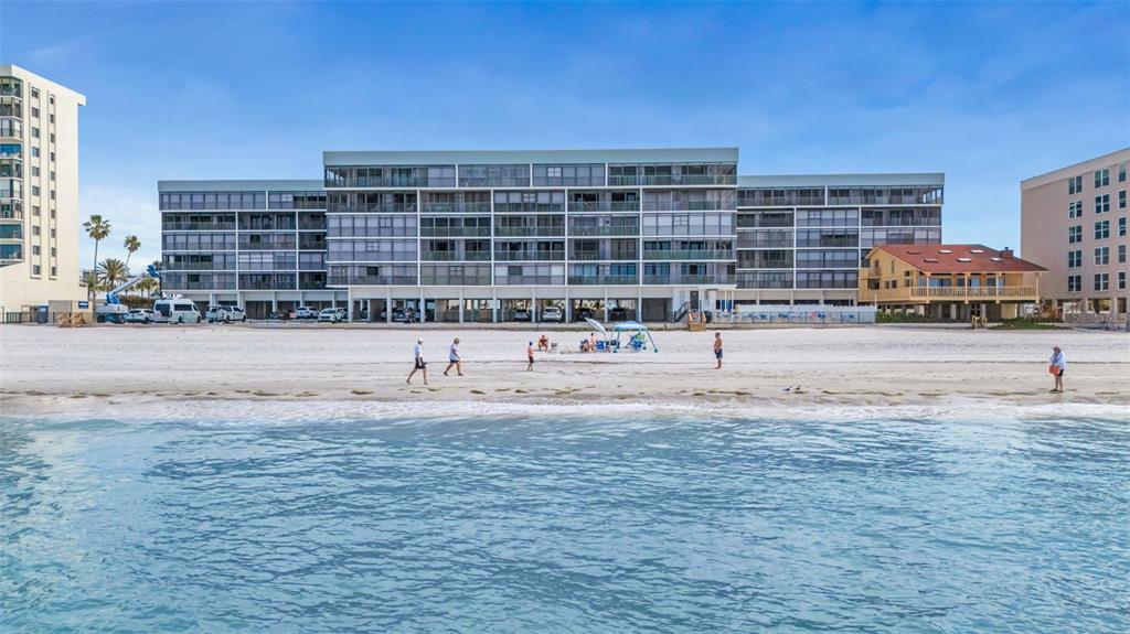 A multi-story beachfront condominium building stands behind a sandy beach with people walking along it — 14900 GULF BOULEVARD UNIT 410, MADEIRA BEACH, FL