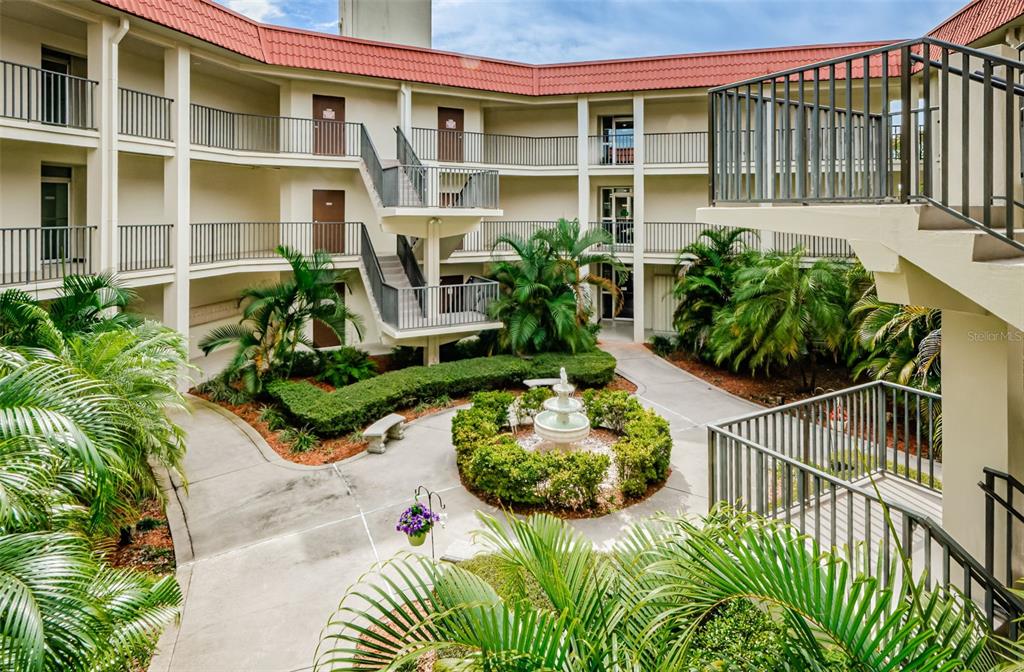 An outdoor courtyard features a central fountain, lush landscaping, and multi-story apartment building walkways — 2700 BAYSHORE BOULEVARD UNIT 2205, DUNEDIN, FL