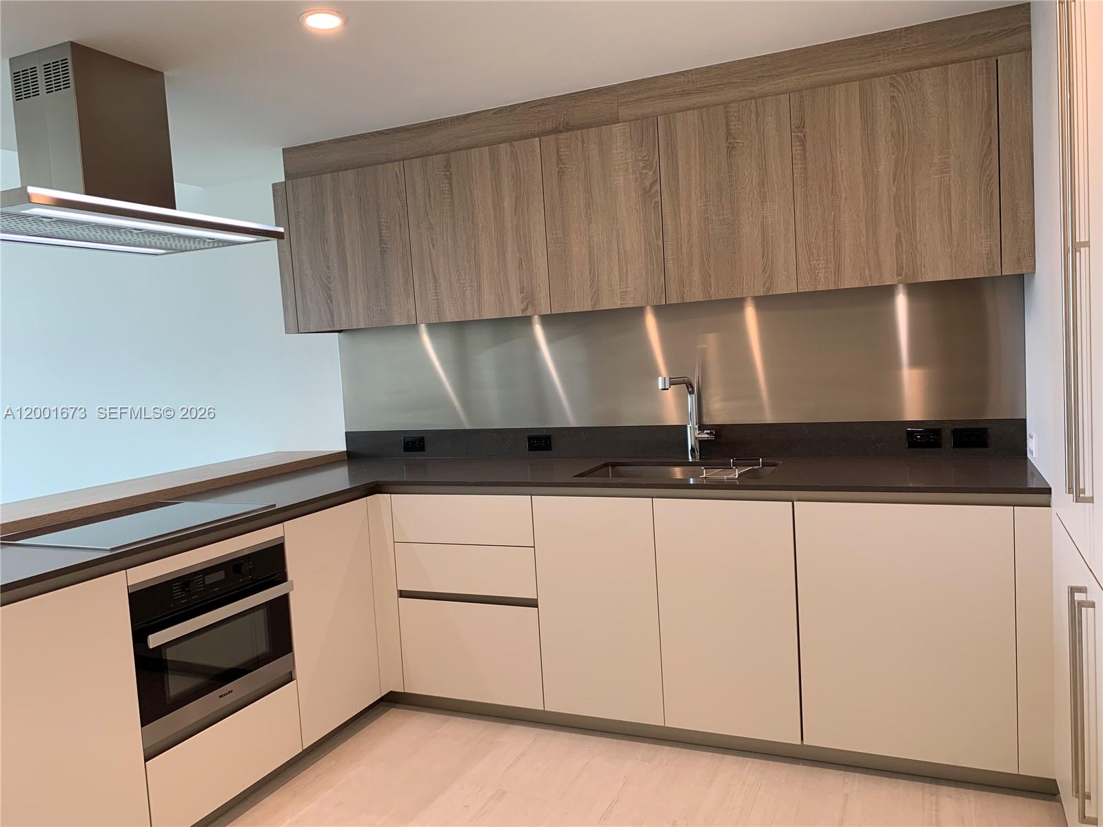 A modern kitchen features light-colored lower cabinets, wood-grain upper cabinets, and a stainless steel backsplash — 1000 Brickell Plz # 3904, Miami FL 33131, MIAMI, FL