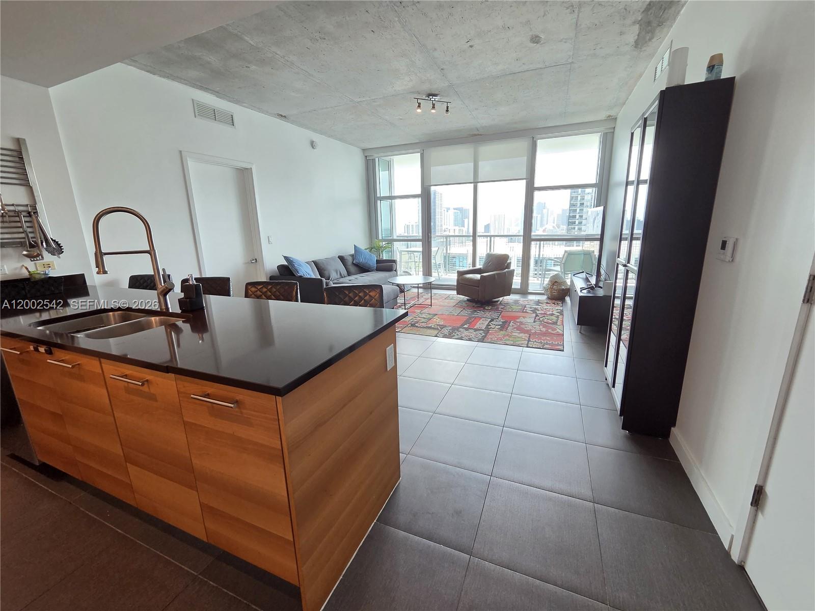 A modern kitchen island with wood cabinets overlooks a living area with floor-to-ceiling windows — 3301 NE 1st Ave # H2710, Miami FL 33137, MIAMI, FL