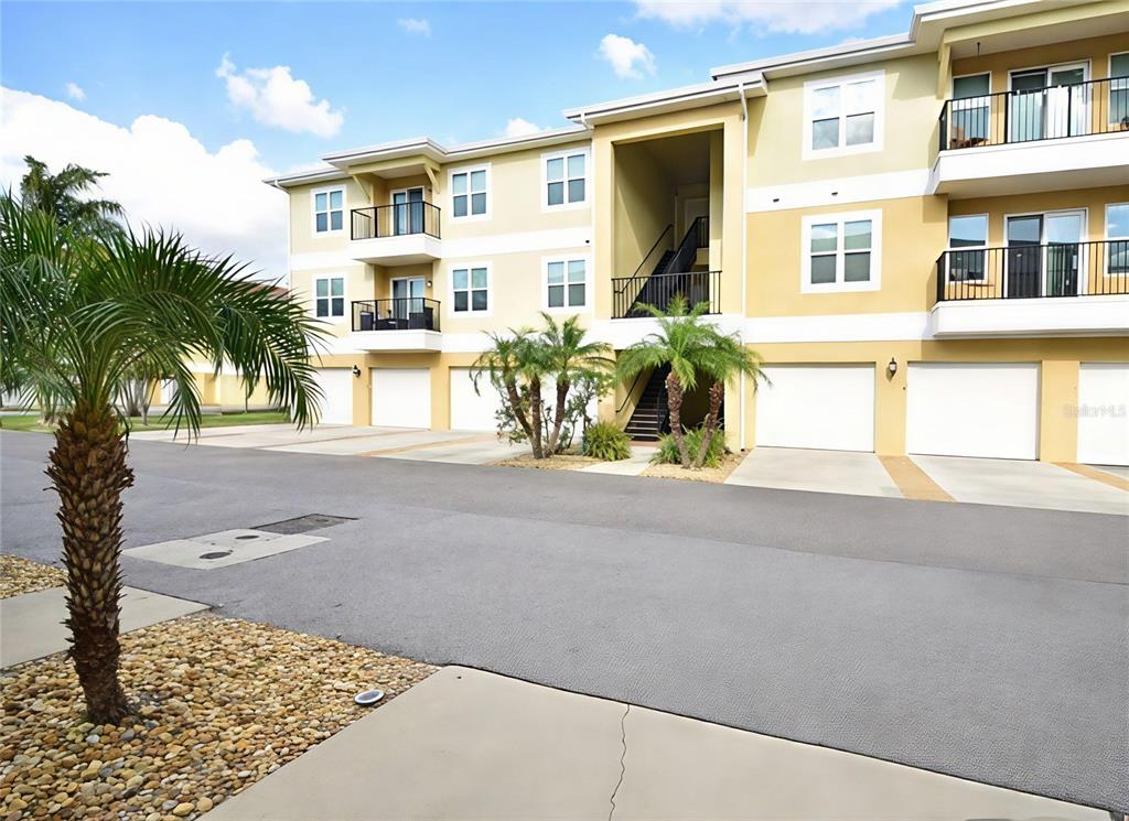 A three-story apartment building features tan stucco walls, white garage doors, and palm trees — 6420 BANYAN BOULEVARD UNIT 303, NEW PORT RICHEY, FL