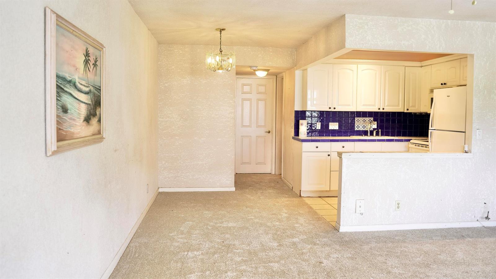 A dining area with beige carpet leads to a kitchen featuring white cabinets and blue tiles — 38 Keswick # B, Deerfield Beach FL 33442, DEERFIELD BEACH, FL