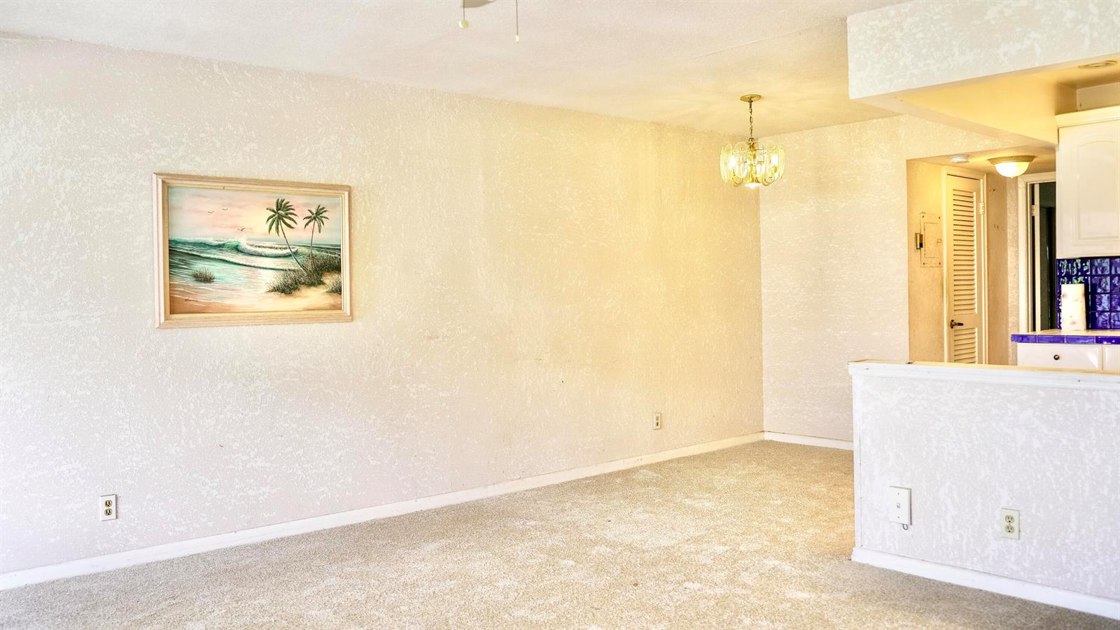 An empty room with beige carpet, textured walls, a hanging chandelier, and a framed painting — 38 Keswick # B, Deerfield Beach FL 33442, DEERFIELD BEACH, FL