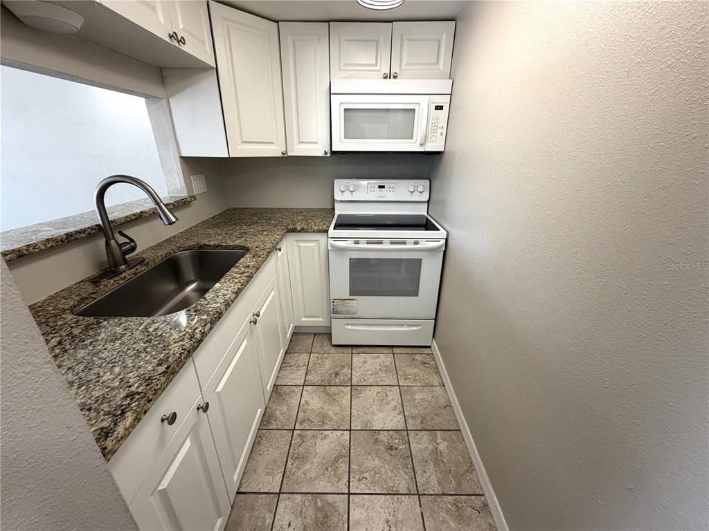 A small kitchen features white cabinets, granite countertops, a stainless steel sink, and white appliances — 7113 E BANK DRIVE UNIT 7113, TAMPA, FL