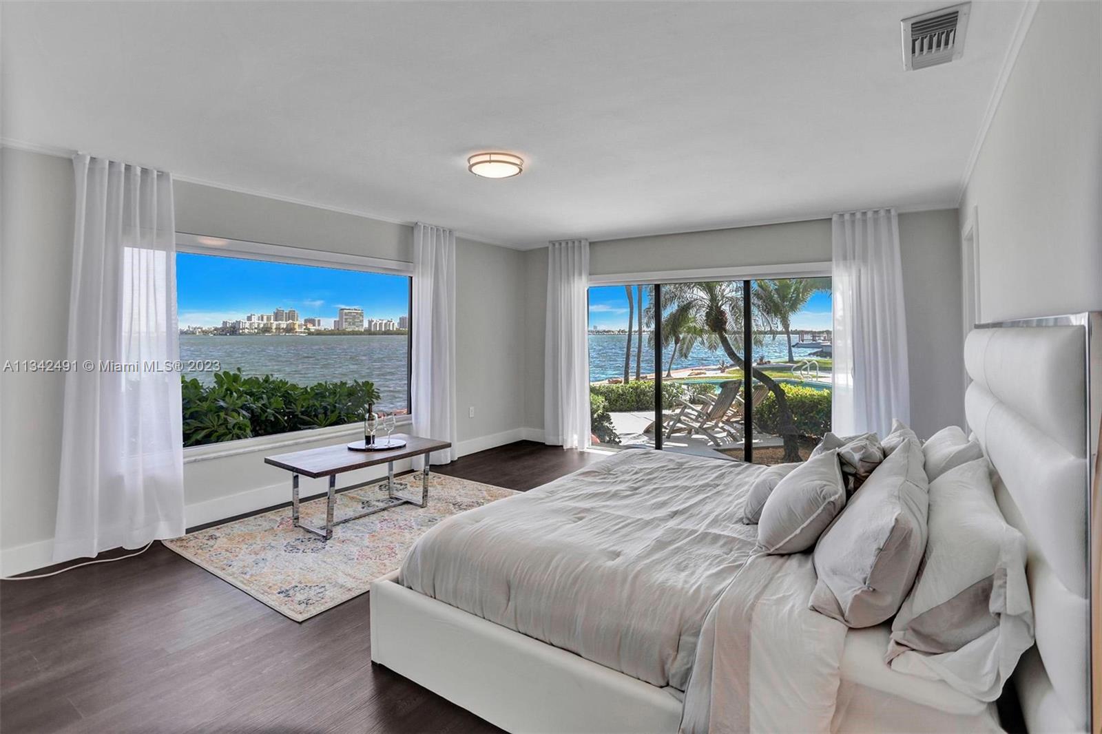 A bedroom features a large bed, dark wood floors, and floor-to-ceiling windows overlooking the water — 9101 N Bayshore Dr, Miami FL 33138, MIAMI, FL