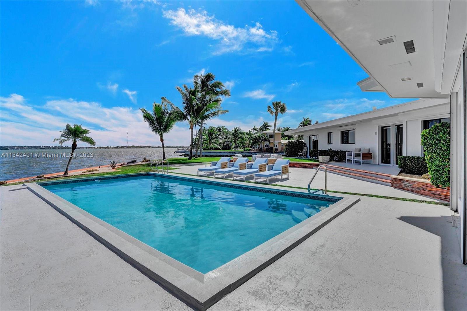 A swimming pool sits on a patio overlooking a body of water with palm trees — 9101 N Bayshore Dr, Miami FL 33138, MIAMI, FL