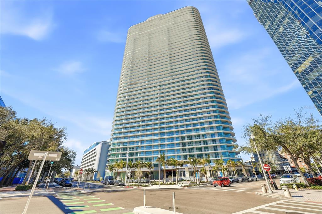 A tall, modern glass residential skyscraper stands at a street intersection on a sunny day — 400 CENTRAL AVENUE UNIT 505, ST PETERSBURG, FL