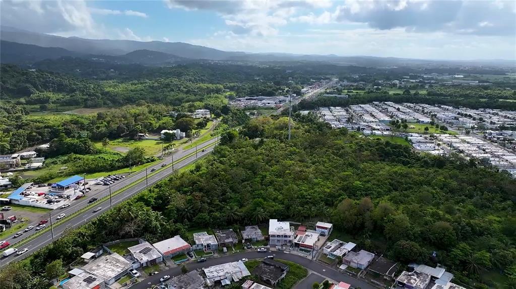 An aerial view shows a residential neighborhood, a highway, and dense forest under a cloudy sky — PR-3 KM 28.5 ZARZAL WARD, RIO GRANDE, PR