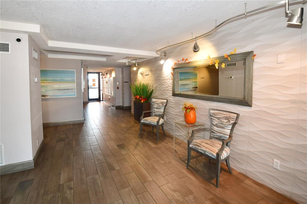 A modern building lobby features wood-look tile floors, textured walls, two chairs, and a mirror — 51 ISLAND WAY UNIT 909, CLEARWATER, FL