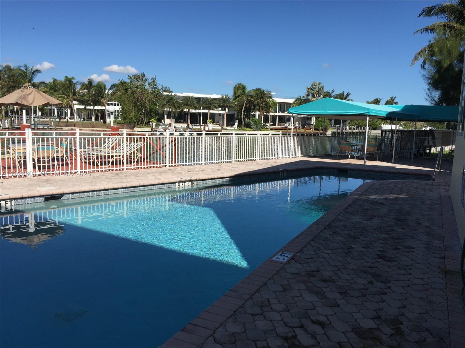 A rectangular swimming pool sits on a paved patio beside a white fence and canal — 4740 Pine Tree Dr # 26, Miami Beach FL 33140, MIAMI BEACH, FL