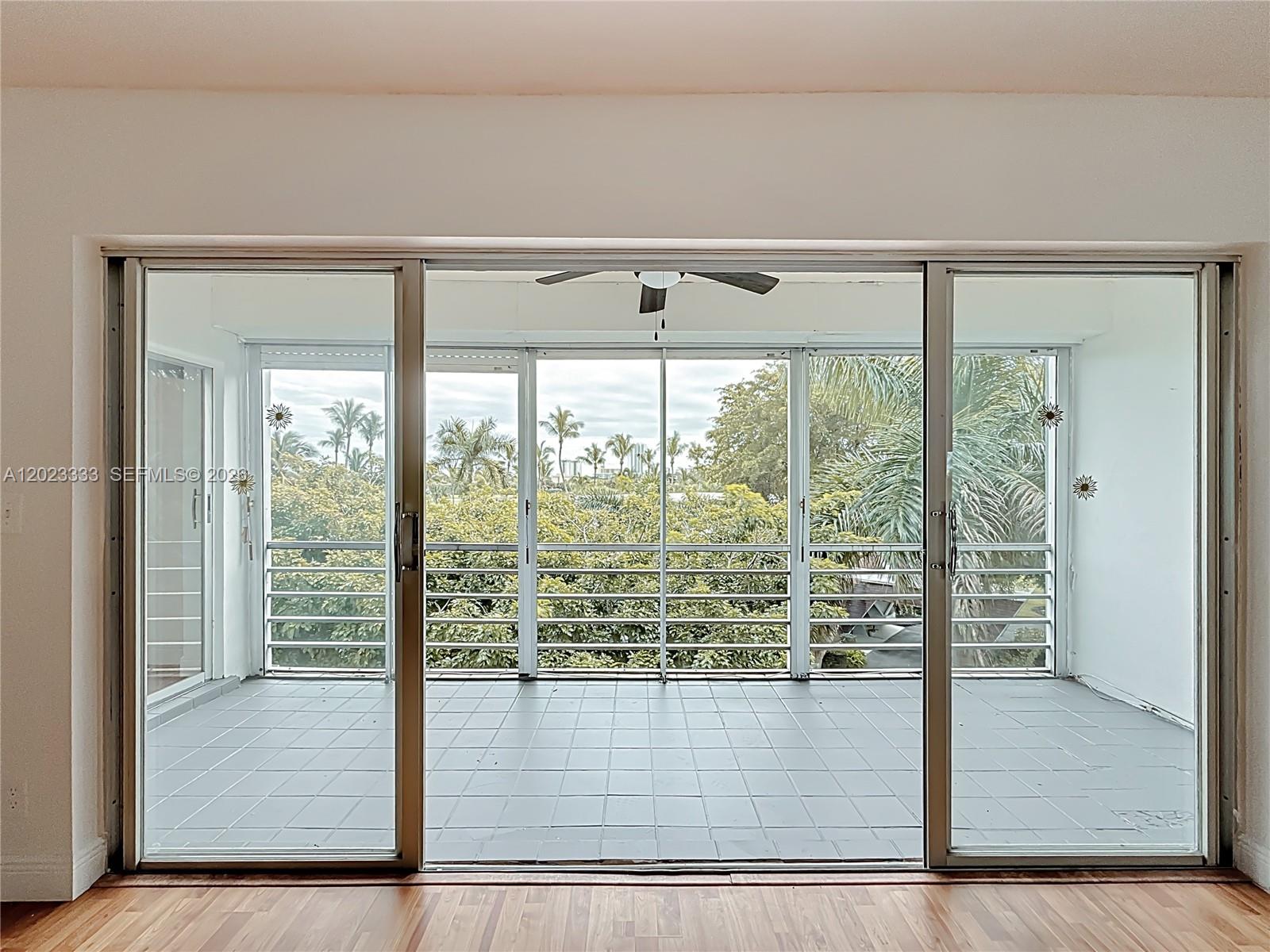 A view through sliding glass doors reveals a tiled balcony overlooking lush green trees — 1000 NE 14th Ave # 411, Hallandale Beach FL 33009, HALLANDALE BEACH, FL