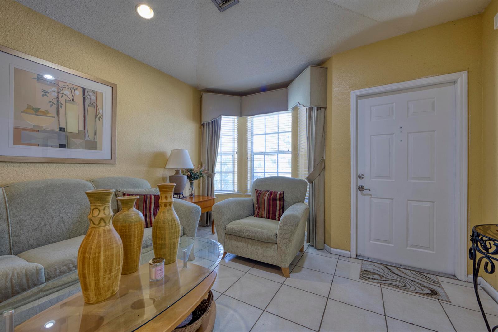 A living room features yellow walls, tile flooring, a glass coffee table, and a bay window — 416 Circle Drive S, Boynton Beach FL 33435, BOYNTON BEACH, FL