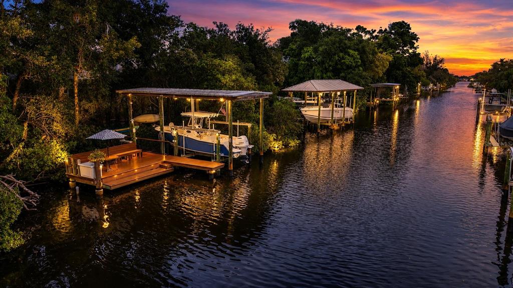 An aerial view of a canal at sunset featuring boat docks, lifts, and a wooden deck — 820 SAND PINE DRIVE NE, ST PETERSBURG, FL