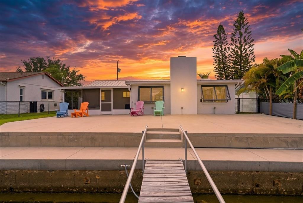 A waterfront house exterior with a concrete patio, four colorful chairs, and a wooden dock — 13902 BARNARD AVENUE, HUDSON, FL