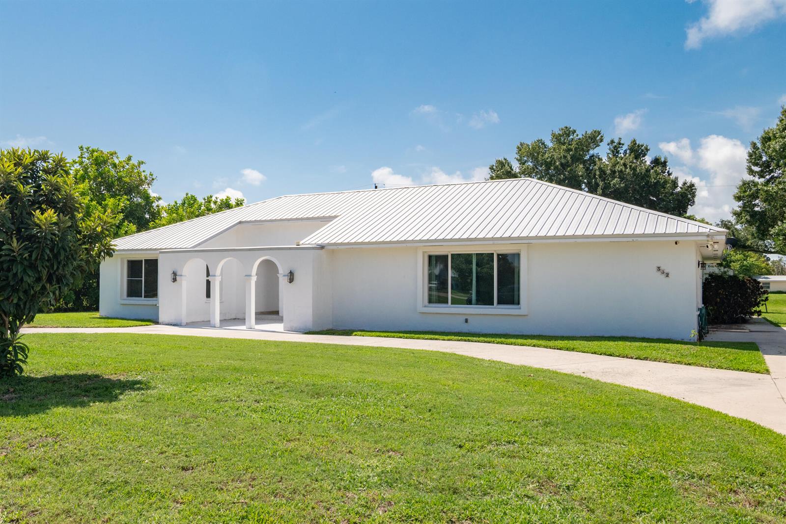 A white single-story house with a metal roof and arched entryway sits on a lawn — 312 De Soto Avenue, Clewiston FL 33440, CLEWISTON, FL