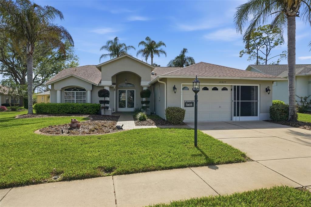 A single-story suburban house with a beige stucco exterior, a two-car garage, and palm trees — 18427 WATER LILY LANE, HUDSON, FL