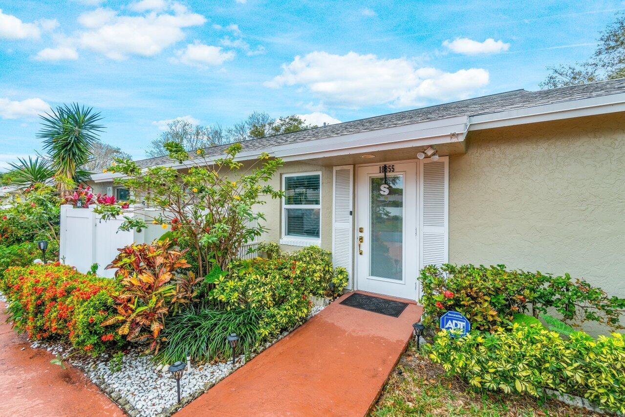 A tan house exterior features a white front door, a red walkway, and lush landscaping — 18855 Candlewick Drive, Boca Raton FL 33496, BOCA RATON, FL