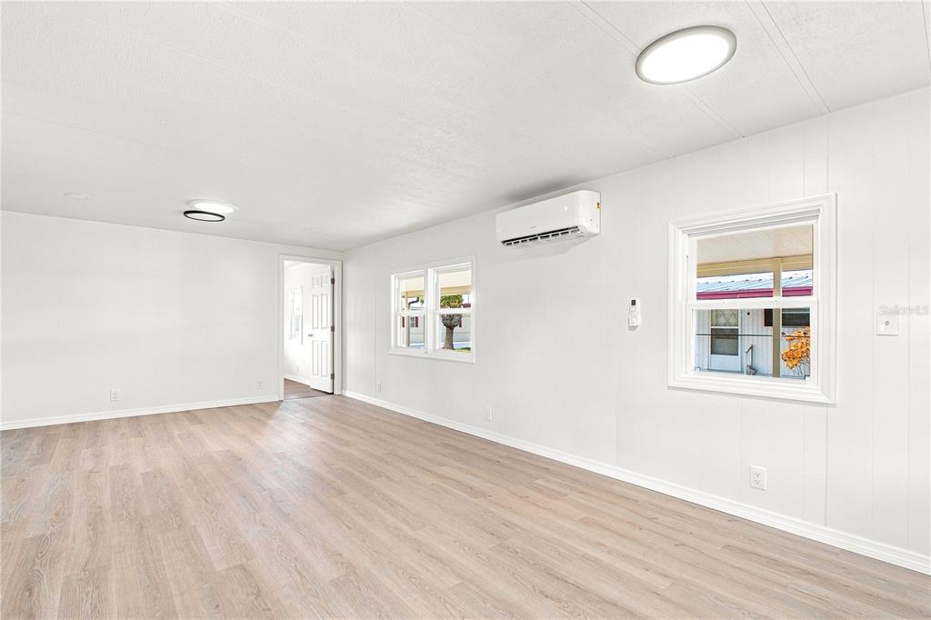 A bright, empty room features white walls, light wood-look flooring, two windows, and a wall-mounted AC — 6711 CLEMENS BOULEVARD, PORT RICHEY, FL