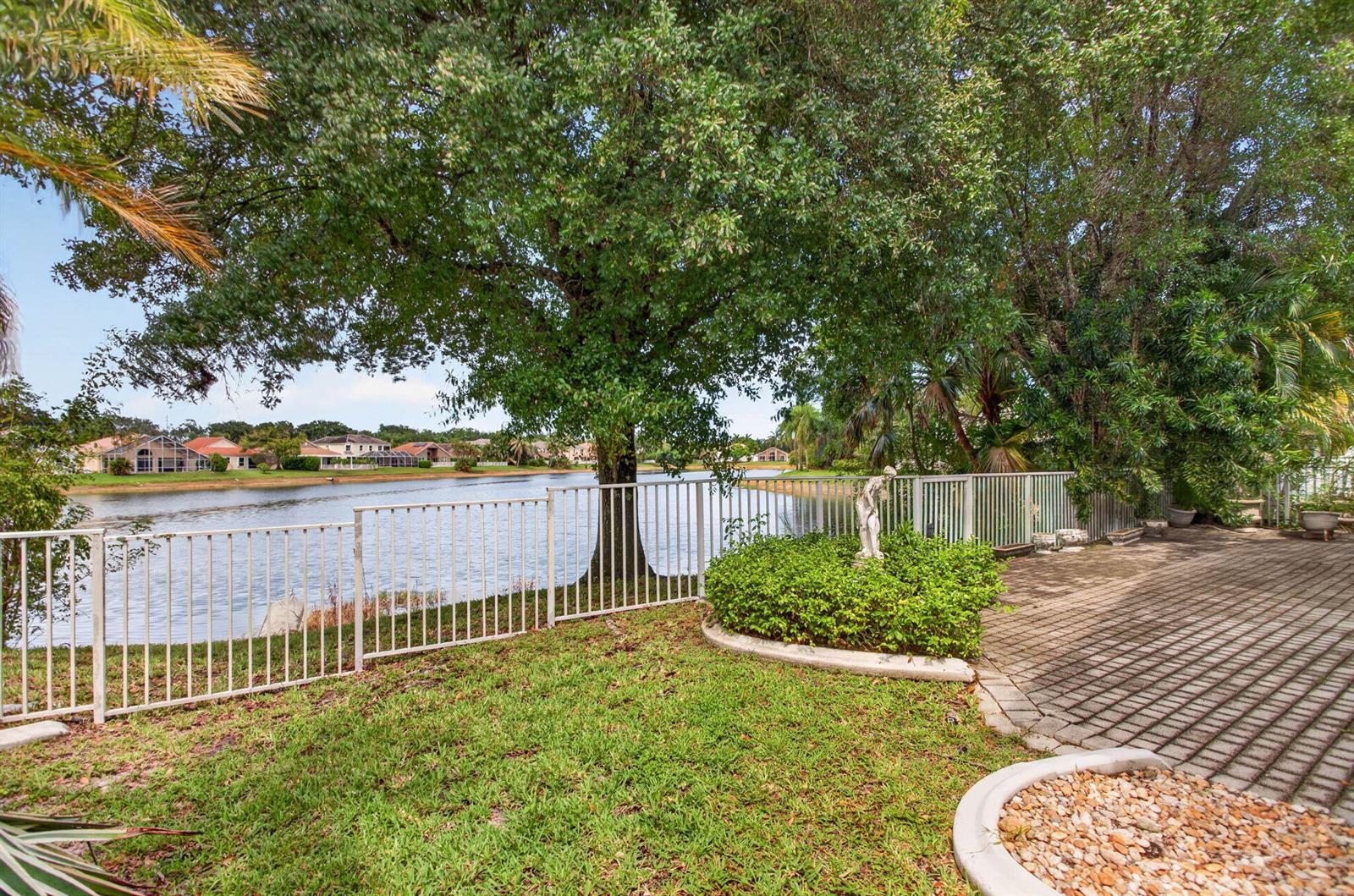 A backyard patio with a white fence overlooks a lake surrounded by trees and houses — 6621 NW 78th Drive, Parkland FL 33067, PARKLAND, FL