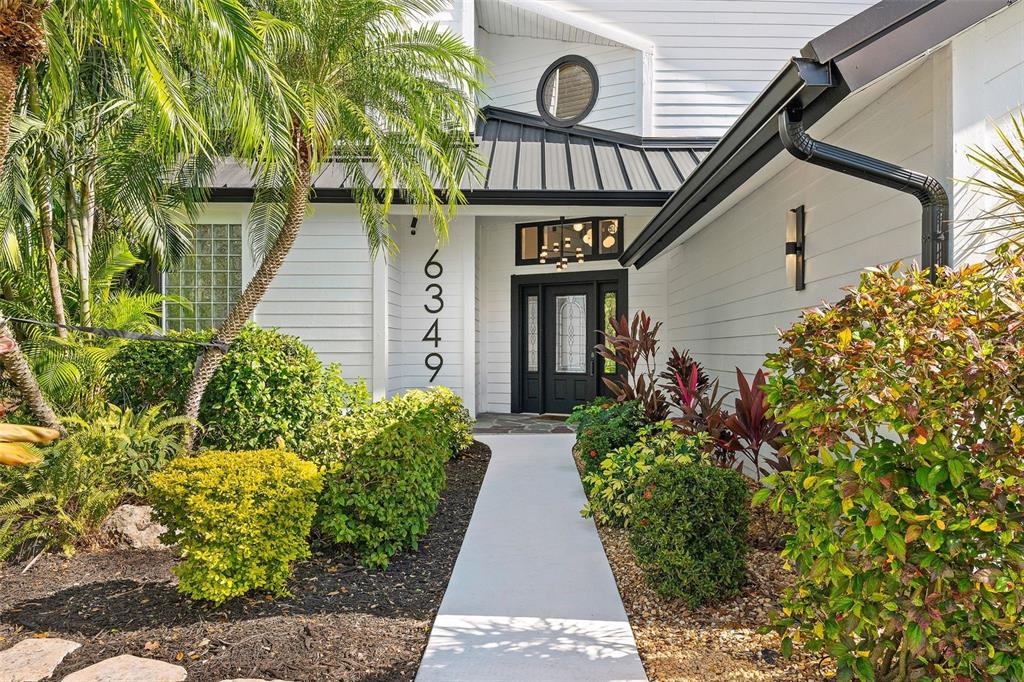 A white house exterior features a black front door, metal roof, and landscaped front yard — 6349 RAVENWOOD WAY, SARASOTA, FL