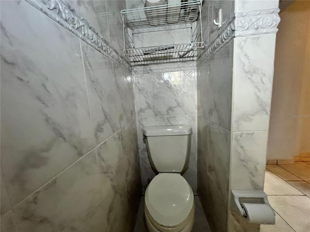 A small toilet room features floor-to-ceiling marble-patterned tile and a wire storage shelf — Urb. Naranjo Valley #45, FAJARDO, PR