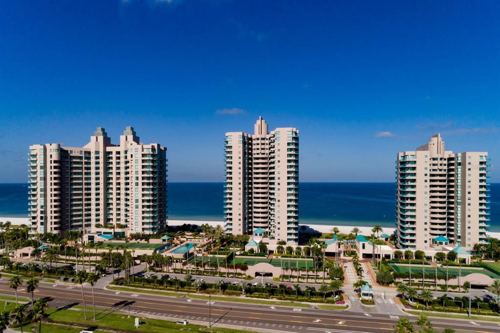An aerial view shows three tall beachfront residential towers with swimming pools and tennis courts — 1560 GULF BOULEVARD UNIT 1605, CLEARWATER BEACH, FL