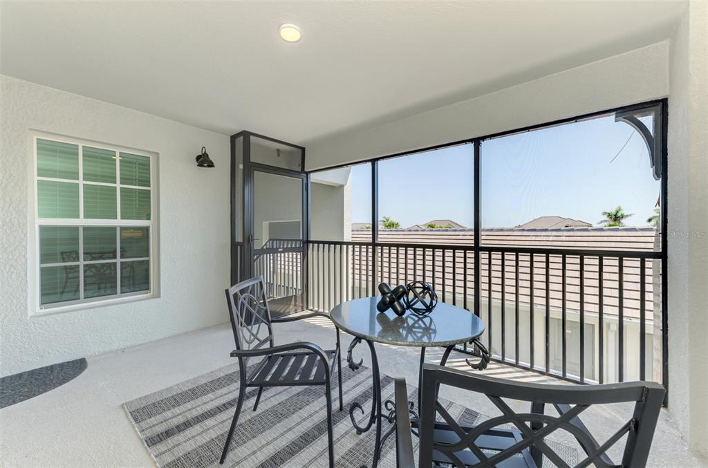 A screened-in balcony features a small bistro set on a rug overlooking a neighborhood — 12470 WELLEN GOLF STREET UNIT 207, VENICE, FL