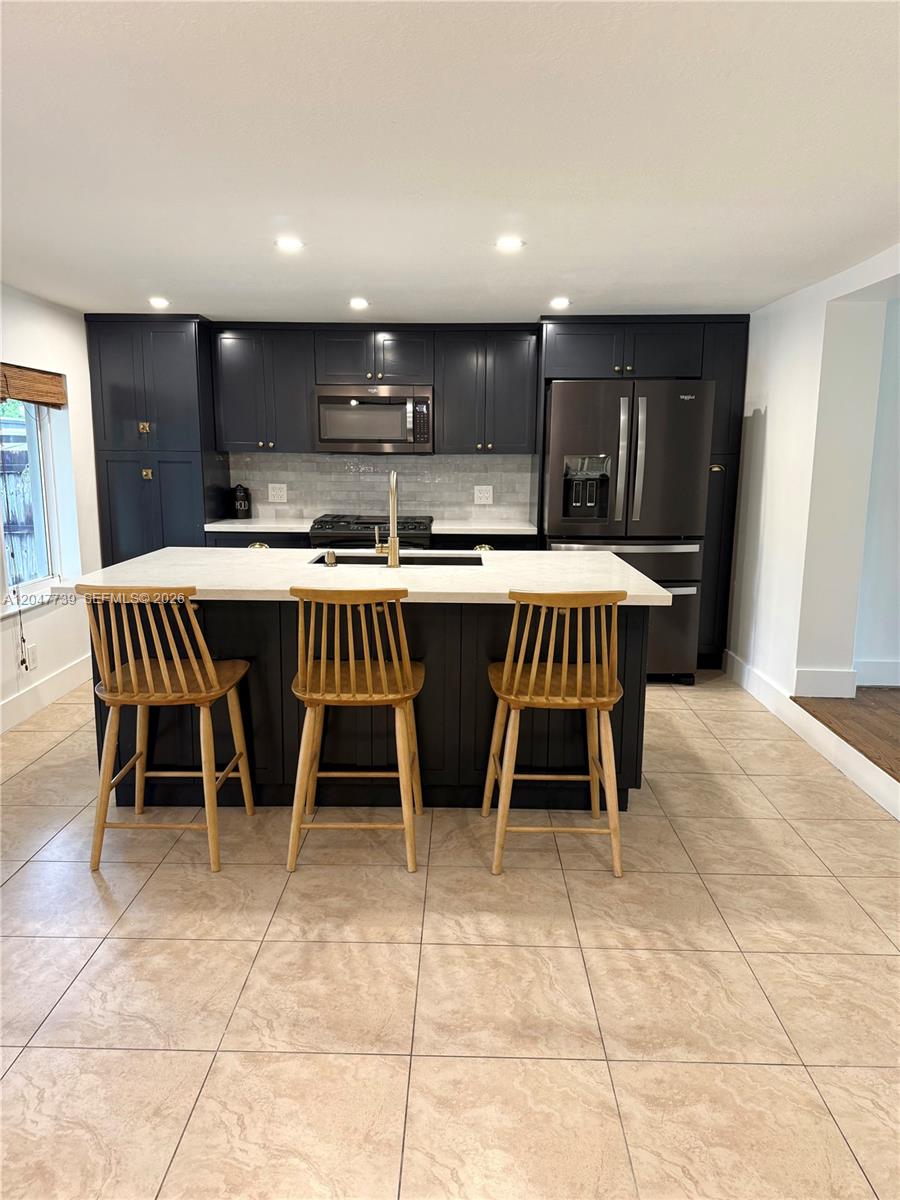 A kitchen features dark cabinetry, a white island with three wooden stools, and tiled flooring — 670 E 58th St, Hialeah FL 33013, HIALEAH, FL