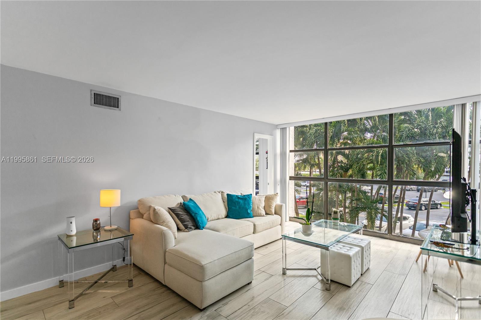 A living room features a beige sectional sofa, glass tables, and large windows overlooking palm trees — 600 Parkview Dr # 204, Hallandale Beach FL 33009, HALLANDALE BEACH, FL