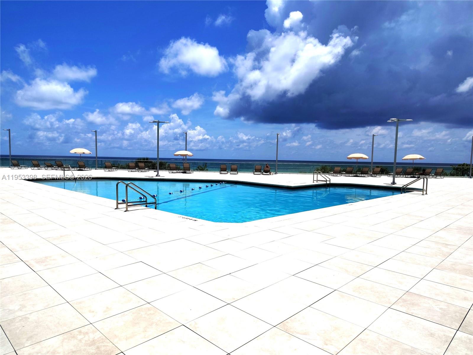 An outdoor swimming pool with lounge chairs overlooks the ocean under a cloudy sky — 7135 Collins Ave # 722, Miami Beach FL 33141, MIAMI BEACH, FL