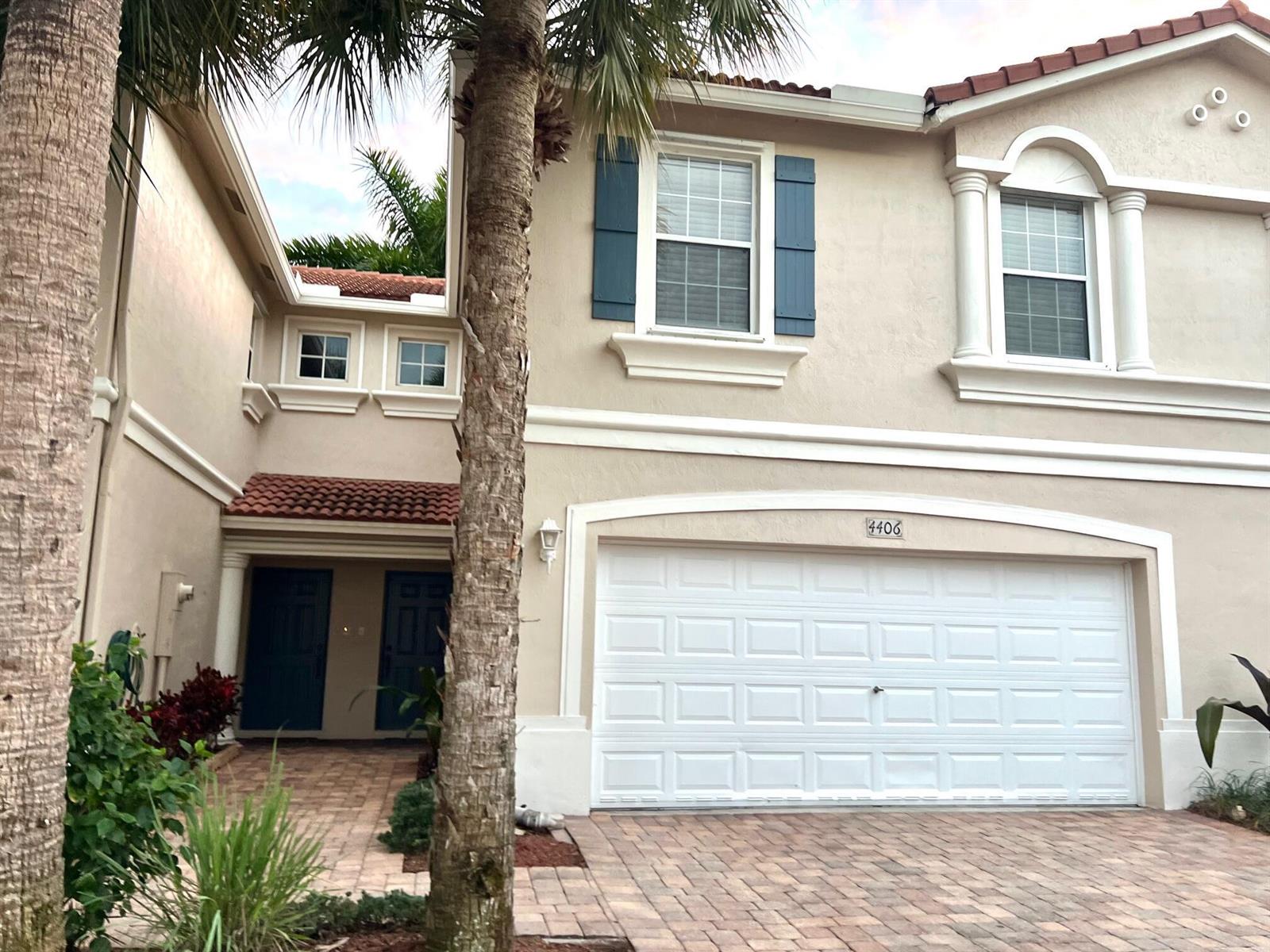 A two-story beige townhouse features a two-car garage, blue shutters, and a brick paver driveway — 4406 NE Windmill Palm Way, FL 33463, FL