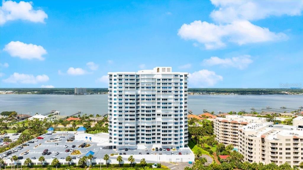 An aerial view shows a tall white residential building surrounded by a parking lot and water — 2 OCEANS WEST BOULEVARD UNIT 702, DAYTONA BEACH, FL