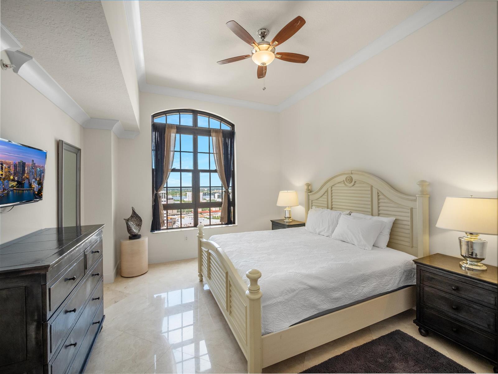 A bedroom with a light-colored bed, dark furniture, tile floors, and a large arched window — 701 S Olive Avenue # 2117, West Palm Beach FL 33401, WEST PALM BEACH, FL