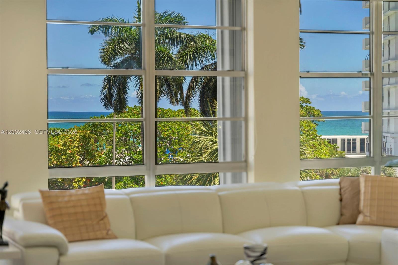 A white sofa sits in a room with large windows overlooking palm trees and the ocean — 5660 Collins Ave # 4B, Miami Beach FL 33140, MIAMI BEACH, FL