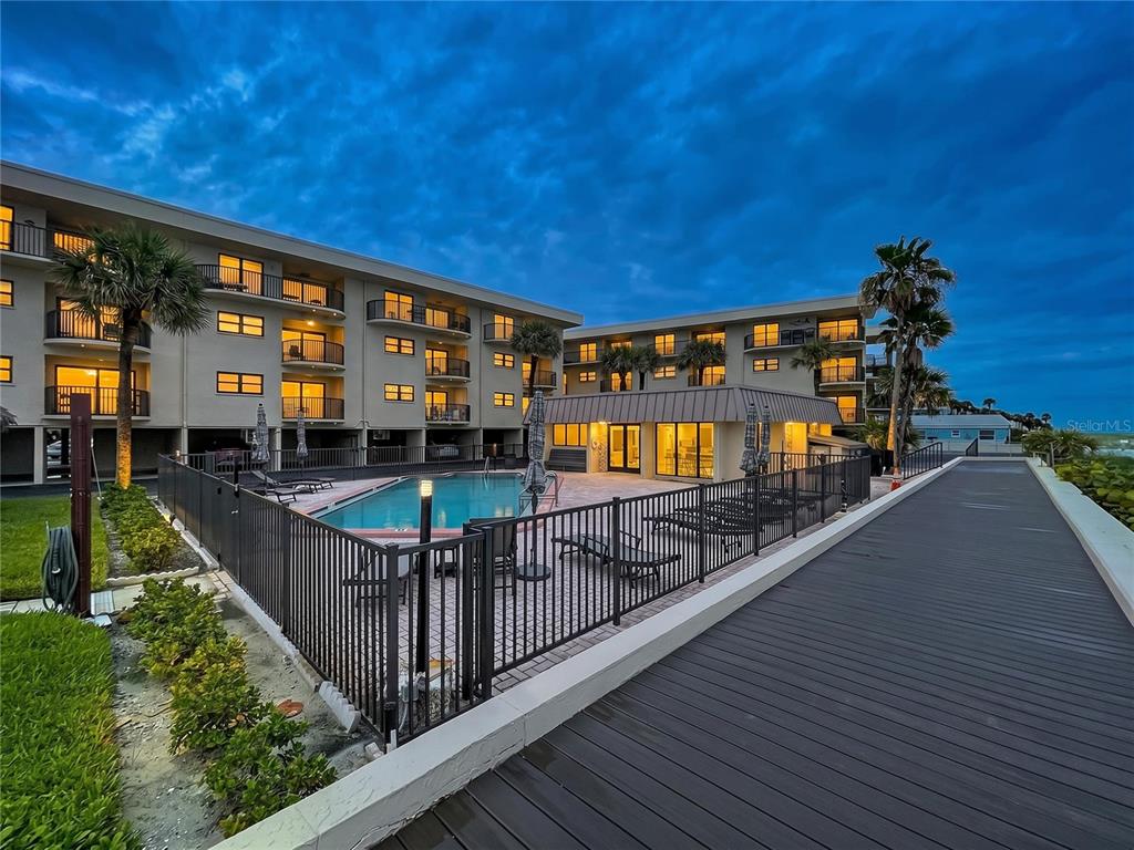 An outdoor swimming pool area with a wooden walkway and multi-story apartment building at dusk — 3500 GULF BOULEVARD UNIT 203, BELLEAIR BEACH, FL