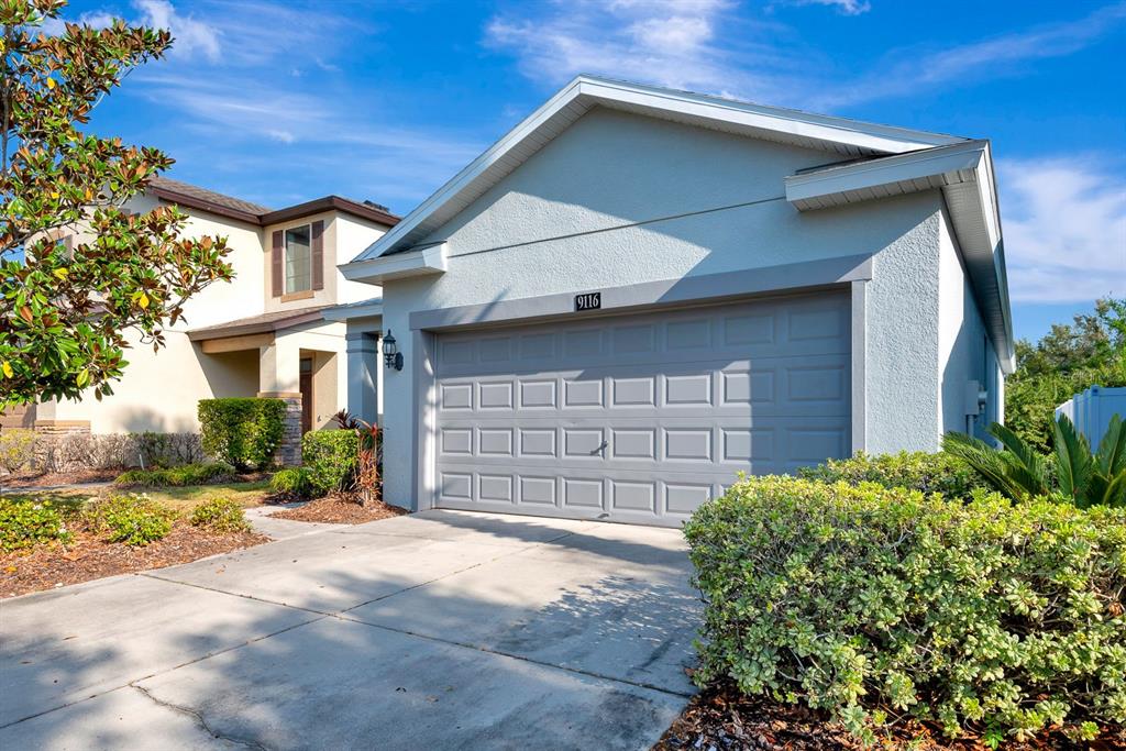 A light-colored house exterior features a gray garage door and a paved driveway — 9116 MOUNTAIN CAMELLIA LANE, RIVERVIEW, FL