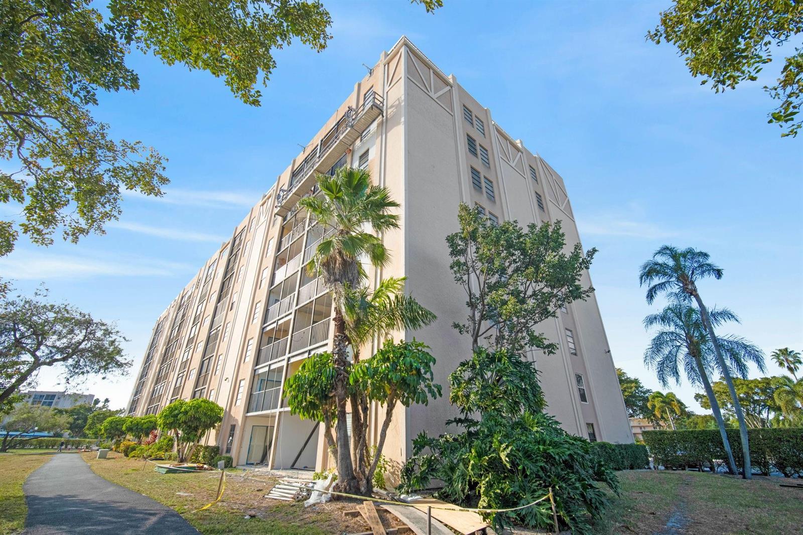 A low-angle exterior view of a multi-story beige apartment building with screened balconies and landscaping — 6866 Huntington Lane # 304, Delray Beach FL 33446, DELRAY BEACH, FL