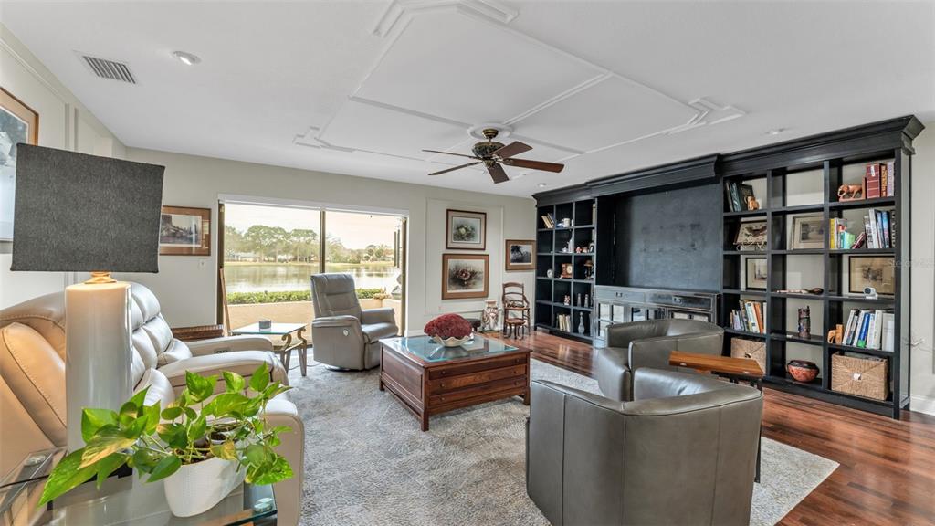A living room features a large built-in black bookcase, a ceiling fan, and lake views — 1007 ROLLING WOODS LANE, LAKELAND, FL