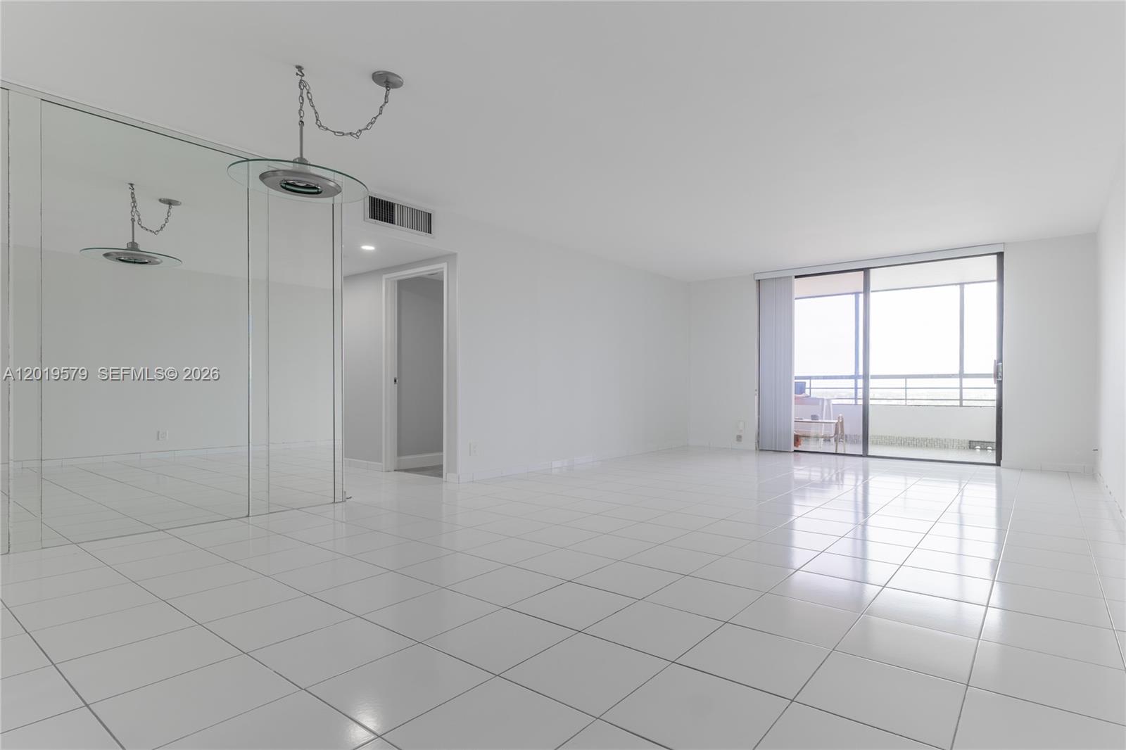 A bright, empty living room features white tile flooring, mirrored walls, and a sliding glass door — 500 Three Islands Blvd # 1110, Hallandale Beach FL 33009, HALLANDALE BEACH, FL