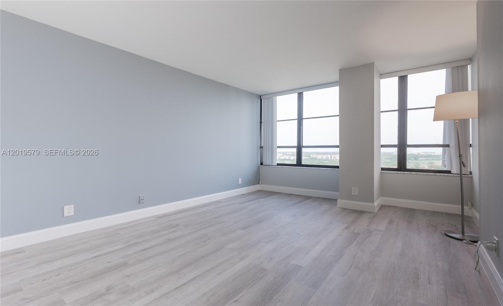 An empty room features light blue walls, gray wood-look flooring, and large windows with views — 500 Three Islands Blvd # 1110, Hallandale Beach FL 33009, HALLANDALE BEACH, FL