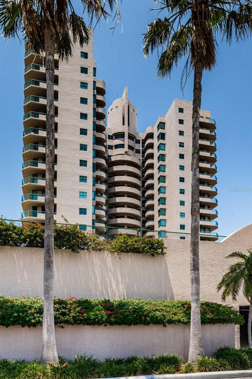A tall, multi-story residential building with curved balconies stands behind a low wall and palm trees — 1520 GULF BOULEVARD UNIT 1101, CLEARWATER BEACH, FL