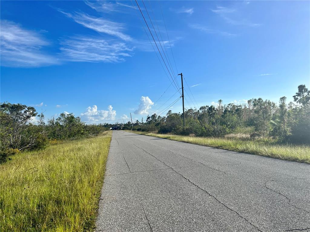 A paved road stretches through a grassy, undeveloped area under a bright blue sky — 9829 SHELBURNE CIRCLE, PORT CHARLOTTE, FL