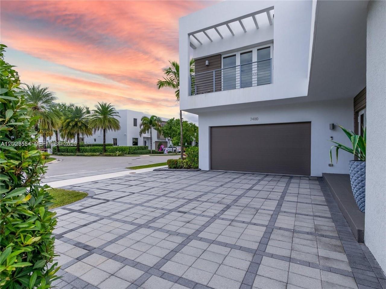 A modern house exterior features a paved driveway, a garage, and a balcony at sunset — 7480 NW 99th Ave, Doral FL 33178, DORAL, FL