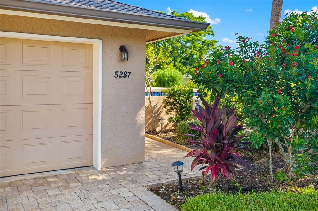 A beige garage door sits beside a paved walkway and a vibrant red-leafed plant — 5287 HERON WAY UNIT 205, SARASOTA, FL